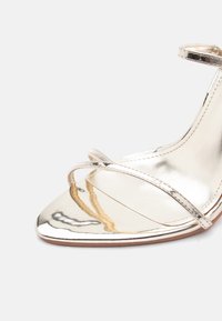 Ασημένιο μεταλλικό πέδιλο με ψηλό τακούνι και ανοιχτή μύτη, με λεπτά λουράκια πάνω από τα δάχτυλα και γύρω από τον αστράγαλο, σε ανοιχτό φόντο.