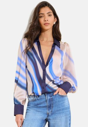 Žena v modro-bielo-čiernej abstraktnej pruhovanej blúzke s veľkými manžetami a svetlomodrých džínsach, ruka neformálne vložená do vrecka.