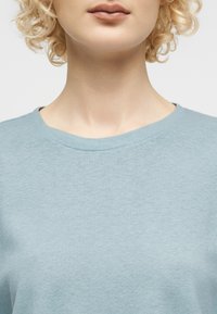 Gros plan d'une personne aux cheveux blonds bouclés portant un t-shirt bleu clair à col rond, montrant le cou et le bas du visage.