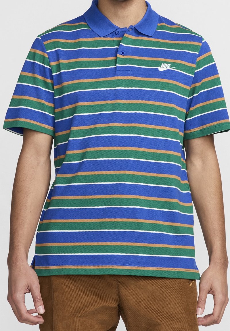 Personne portant un polo Nike à rayures bleues, vertes et dorées avec un col bleu, debout devant un fond uni. Buste visible.