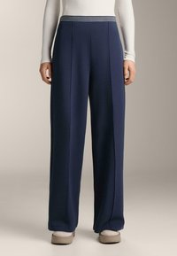 Osoba oblečená v námorníckych širokých nohaviciach s pruhovaným pásom, bielom tričku s dlhým rukávom, bielych ponožkách a béžových nazúvacích topánkach.