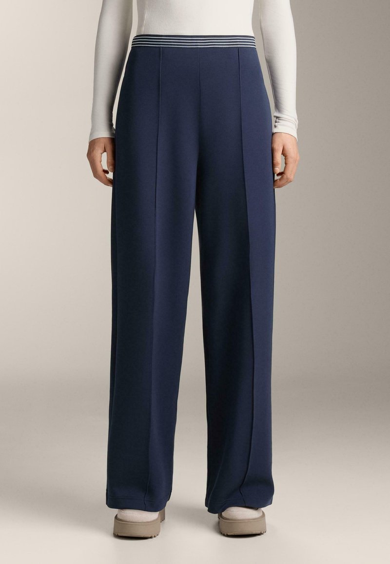 Osoba oblečená v námorníckych širokých nohaviciach s pruhovaným pásom, bielom tričku s dlhým rukávom, bielych ponožkách a béžových nazúvacích topánkach.