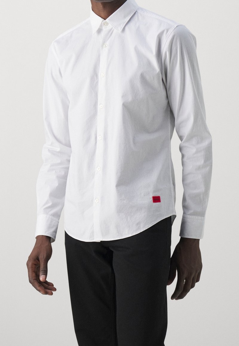 Homme portant une chemise blanche à manches longues avec une petite étiquette rouge en bas devant, associée à un pantalon noir.