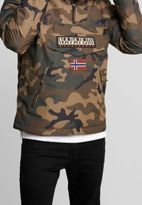 Chaqueta con capucha de patrón de camuflaje en tonos terrosos, que incluye un bolsillo con cremallera, parche con logo y acento de la bandera de Noruega. Textura suave.
