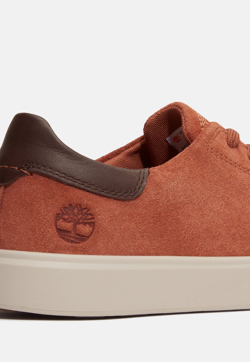 Zapatillas de ante en color naranja oxidado, con talón de cuero marrón, logo grabado y suela de goma blanca. Textura suave con detalles cosidos.