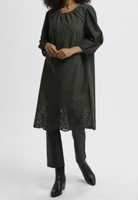 Vestido túnica verde oscuro con cuello redondo, detalle de ojal y acentos de encaje calado en las mangas y el dobladillo. Combinado con pantalones negros y botas.