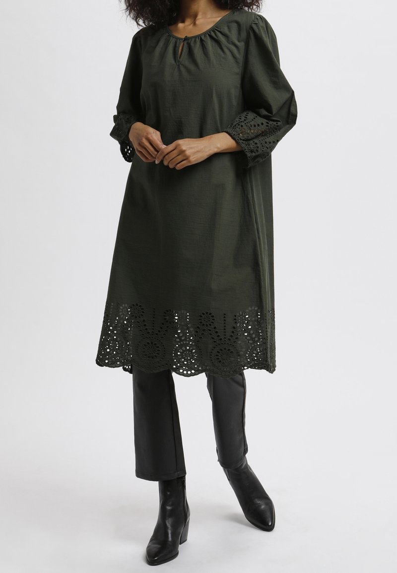 Vestido túnica verde oscuro con cuello redondo, detalle de ojal y acentos de encaje calado en las mangas y el dobladillo. Combinado con pantalones negros y botas.
