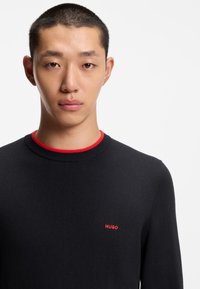Maglione nero con colletto rotondo rosso e piccolo logo "HUGO" rosso sul petto. Il tessuto è liscio con un design aderente.