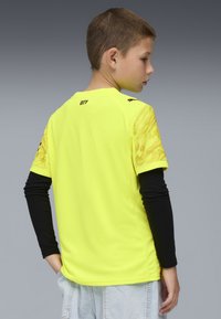 T-shirt atletico giallo brillante con maniche corte a fantasia, maglietta a maniche lunghe nera visibile sotto, e colletto rotondo.