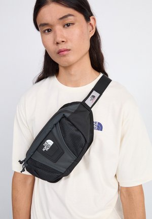 Person iført en hvid The North Face t-shirt og en sort og grå The North Face crossbody taske på en lys baggrund.