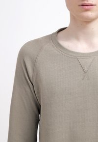 Khaki langetermijnshirt met ronde hals en raglanmouwen. Heeft een geribbelde textuur en een subtiele gestikte detail op de kraag.