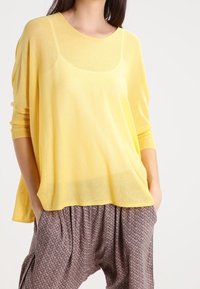 Gelber leichter Pullover mit weitem Ausschnitt und Dreiviertelärmeln, kombiniert mit gemusterten, locker sitzenden Hosen. Strukturiertes Material.