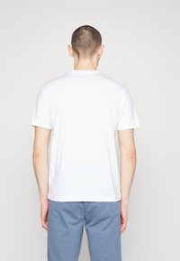 Mann mit kurzen Haaren, der ein einfaches weißes T-Shirt und blaue Hosen trägt, steht mit dem Rücken zur Kamera vor einem schlichten Hintergrund.
