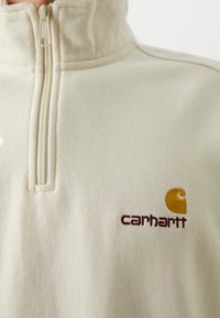 Pull-over en polaire crème avec un col montant, doté d'une fermeture à demi-zip, et logo "Carhartt" brodé avec des accents marron et jaune.