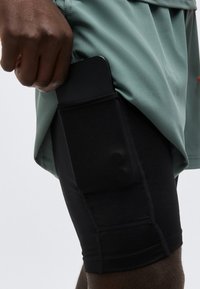 Χέρι που τοποθετεί ένα smartphone σε μαύρη υφασμάτινη τσέπη στο πλάι μαύρων εφαρμοστών σορτς, φορώντας πράσινο πουκάμισο.