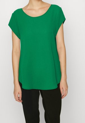 Woman wearing a loose-fitting, short-sleeve green top with a round neckline and black pants, shown from shoulders to hips.