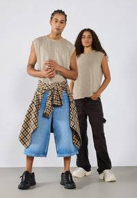 Beige ermeless skjorter, blå vide jeans med rutete skjorte knyttet i livet. Svart og hvit sneakers med teksturerte overflater.