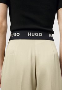 Czarny pas z logo "HUGO" w metalicznym napisie; zestawiony z plisowanymi beżowymi spodniami wykonanymi z gładkiego materiału.