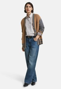 Cardigan en tricot marron et gris avec des boutons, accents rayés, superposé à une chemise à rayures claire. Porté avec un jean ample bleu et des chaussures noires.