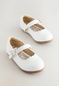 Zapatos de niña pequeños blancos tipo Mary Jane con plantillas florales, bordes en forma de concha y cierres de mariposa sobre un fondo claro.