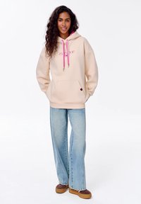Béžová oversized mikina s růžovými stahovacími šňůrkami a přední kapsou, kombinovaná se světle modrými širokými džíny a hnědými botami, stojící na jednobarevném pozadí.