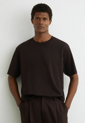 Young man with short curly hair wearing a plain dark brown T-shirt and matching pants, standing with hands in pockets against white wall.