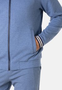 Veste zippée bleue avec poches latérales, poignets rayés contrastés, texture douce et coupe décontractée. Le matériau semble léger et respirant.