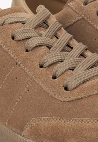 Brown suede sneakers feature a textured surface, stitched accents, and flat beige laces woven through reinforced eyelets.