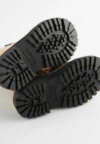 Schwarze Gummisohlen mit tiefen Profilen und strukturierten Oberflächen; die "NEXT"-Branding ist auf den Sohlen sichtbar. Insgesamt robustes und funktionales Design.