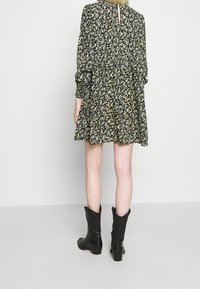 Svart klänning med blommönster, långärmad, med sammanknuten midja och knälång fåll; tillsammans med svarta ankelskor. Bakifrån visad.