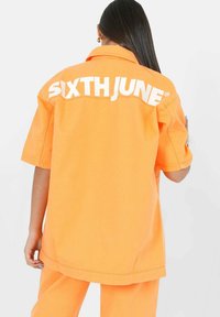 Camisa oversized naranja de denim, con un logo en la parte posterior que dice "SIXTHJUNE" en blanco. Mangas cortas con un ajuste relajado y cuello tipo polo.