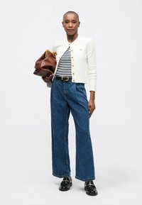 Mujer vistiendo un cárdigan blanco abotonado sobre una camiseta de rayas, jeans de pierna ancha de talle alto, mocasines negros y sosteniendo un gran bolso de cuero marrón.