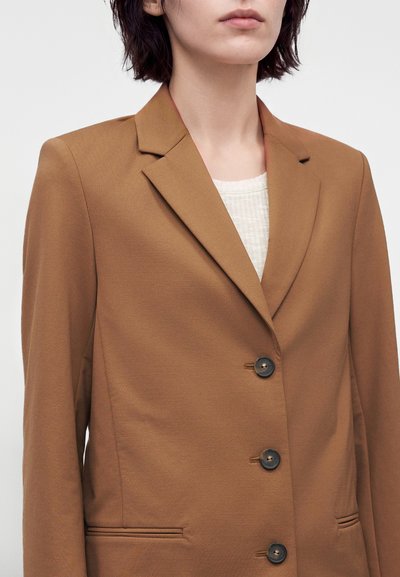 Femme portant un blazer marron à trois boutons noirs sur un top blanc texturé, montrée du cou aux hanches sur un fond uni.