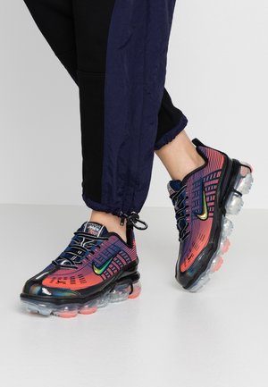 Personne portant un pantalon de jogging noir et bleu marine avec des baskets Nike Vapormax violettes et orange, dotées de semelles translucides.