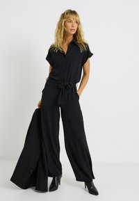 Jumpsuit noir à manches courtes, boutons sur le devant, taille ceinturée, design à larges jambes et tissu texturé ; associé à un blazer noir et des bottes à talons.