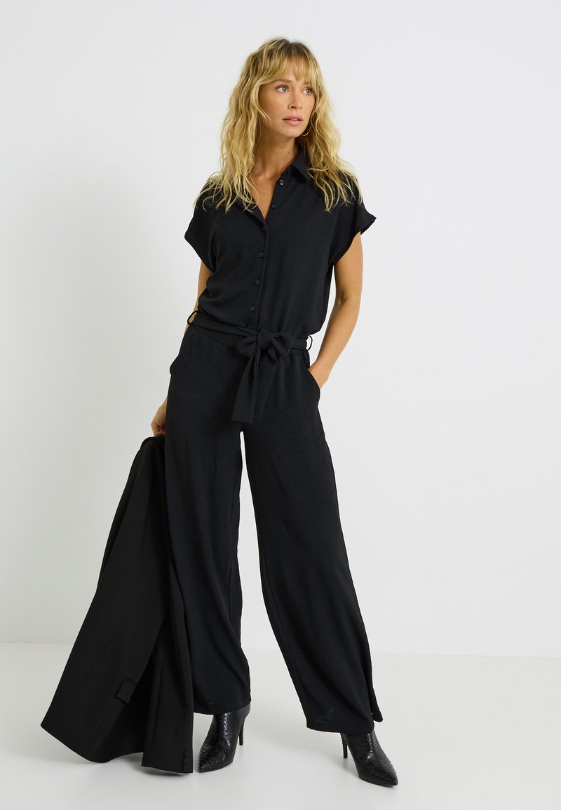 Jumpsuit noir à manches courtes, boutons sur le devant, taille ceinturée, design à larges jambes et tissu texturé ; associé à un blazer noir et des bottes à talons.