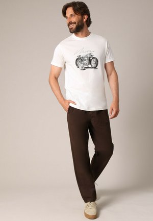 Witte katoenen T-shirt met motorgraphic, gecombineerd met donkerbruine zachte stoffen broek en lichtbeige sneakers. Casual outfit.
