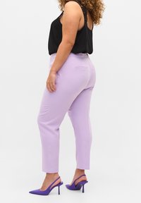 Kvinna som bär skräddarsydda lilafärgade byxor, svart ärmlös topp med spetsdetalj och lila högklackade slingback-skor, stående i profil.