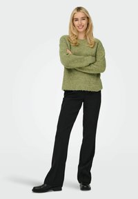 Groene gebreide trui met ronde hals, gecombineerd met zwarte rechte broek en zwarte schoenen. Model staat met de armen gekruist.