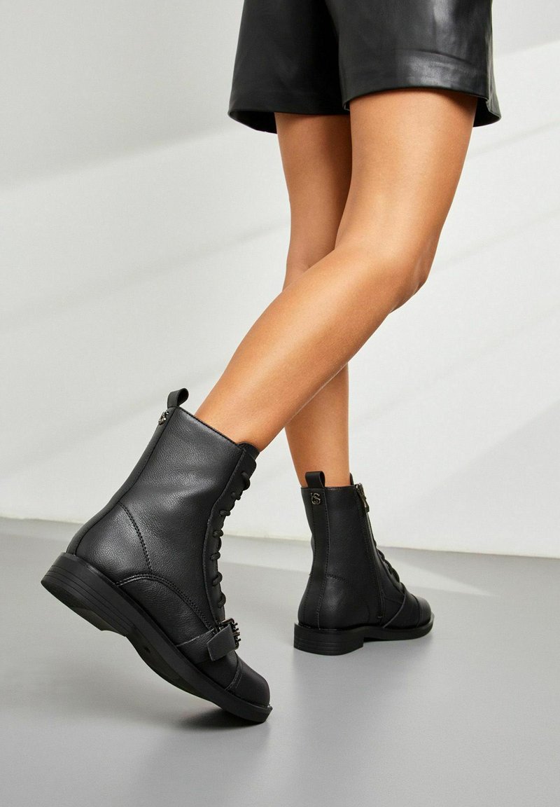 Des bottines en cuir noir présentent un bout arrondi, des lacets à l'avant, une fermeture éclair latérale et une petite boucle décorative au niveau de la cheville. Semelle en caoutchouc solide.