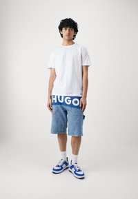 Camiseta de algodón blanca, pantalones cortos de mezclilla azules con el logo de HUGO y zapatillas azules y blancas. Los pantalones cortos tienen un corte relajado y costuras visibles.