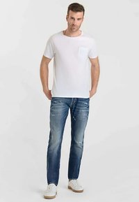 Vit t-shirt med korta ärmar och en bröstficka, kombinerad med blekta blå jeans och vita sneakers med beige sula, stående mot en enfärgad bakgrund.