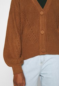 Brun stickad cardigan med djup V-ringning, tre stora knappar och puffärmar. Har texturerade mönster och ribbad fåll.