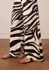 Zwart-witte zebra gestreepte wijde broek met gouden accenten, gecombineerd met metallic zilveren platte sandaal, op een houten vloer.