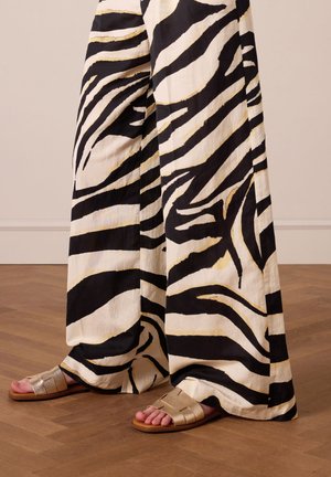 Zwart-witte zebra gestreepte wijde broek met gouden accenten, gecombineerd met metallic zilveren platte sandaal, op een houten vloer.