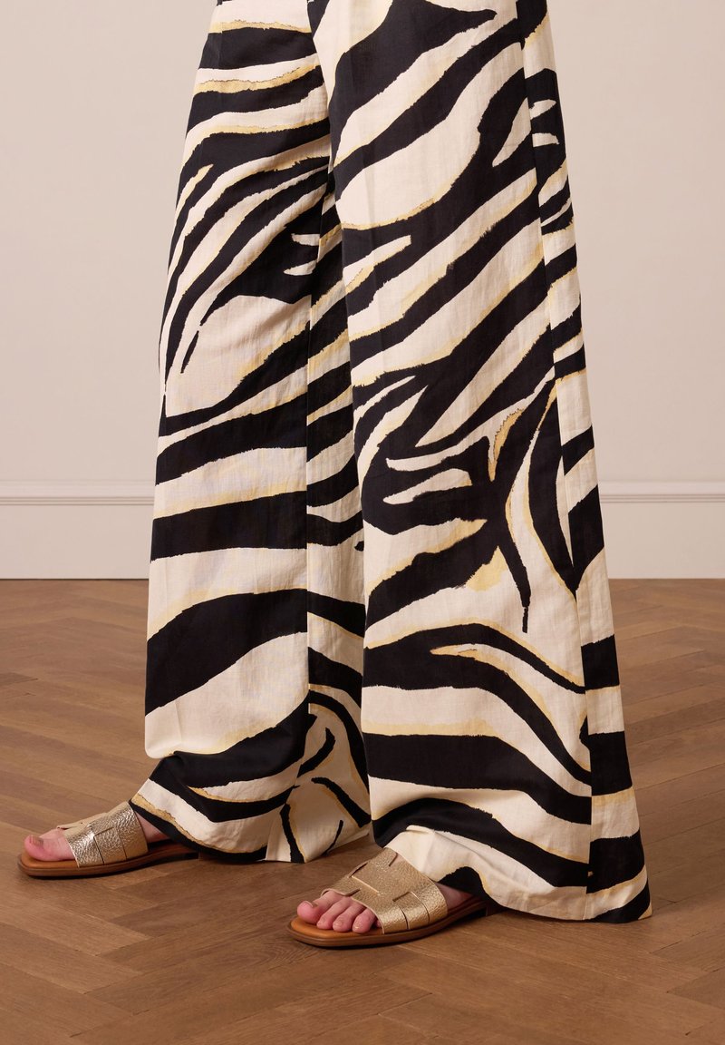 Zwart-witte zebra gestreepte wijde broek met gouden accenten, gecombineerd met metallic zilveren platte sandaal, op een houten vloer.