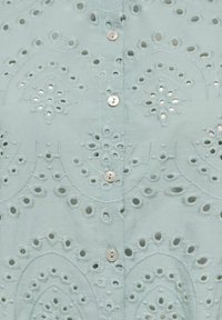 Tissu bleu clair avec une broderie œil-de-perdrix circulaire et florale complexe, et de petits boutons blancs alignés verticalement.