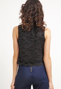Top corto in pizzo nero con motivo floreale, orlo a scallop e senza maniche. Indossato sopra pantaloni a vita alta blu, mostrando dettagli testurizzati.