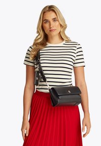 Νεαρή γυναίκα με μακριά ξανθά μαλλιά, που φοράει ένα μαύρο-άσπρο ριγέ πουκάμισο, κόκκινη πλισέ φούστα και μια μαύρη τσάντα χιαστί με θήκη.