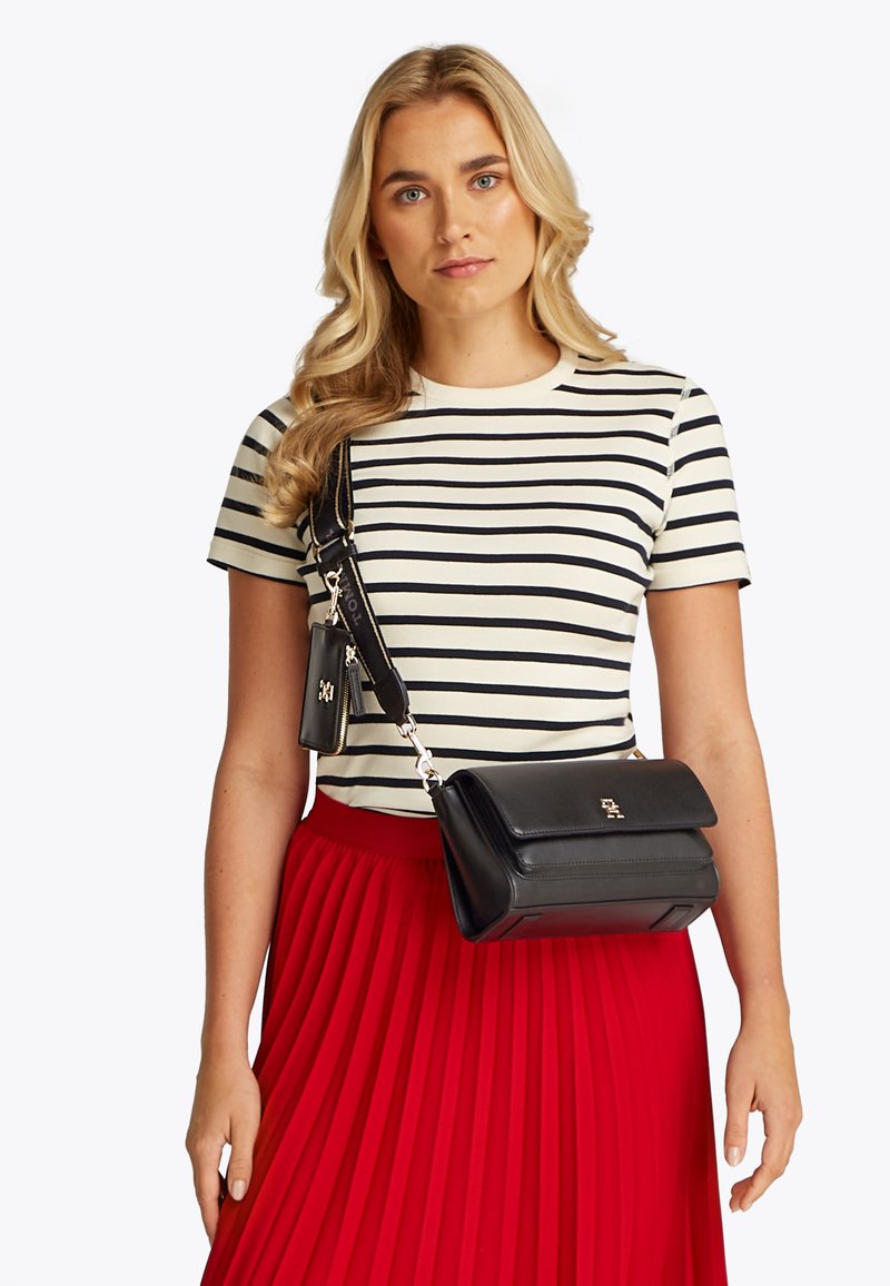 Νεαρή γυναίκα με μακριά ξανθά μαλλιά, που φοράει ένα μαύρο-άσπρο ριγέ πουκάμισο, κόκκινη πλισέ φούστα και μια μαύρη τσάντα χιαστί με θήκη.